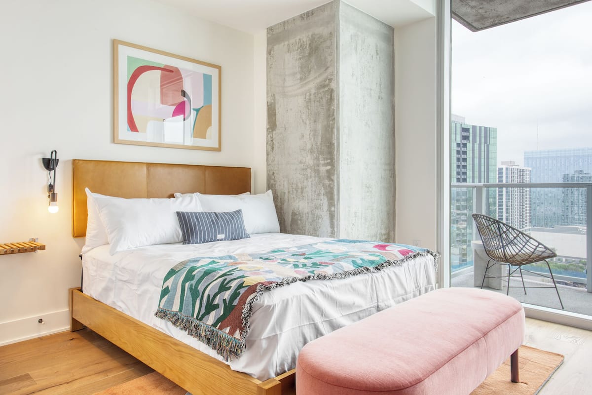 A bedroom with a large bed, a pink ottoman, a chair by the window, and a city view.