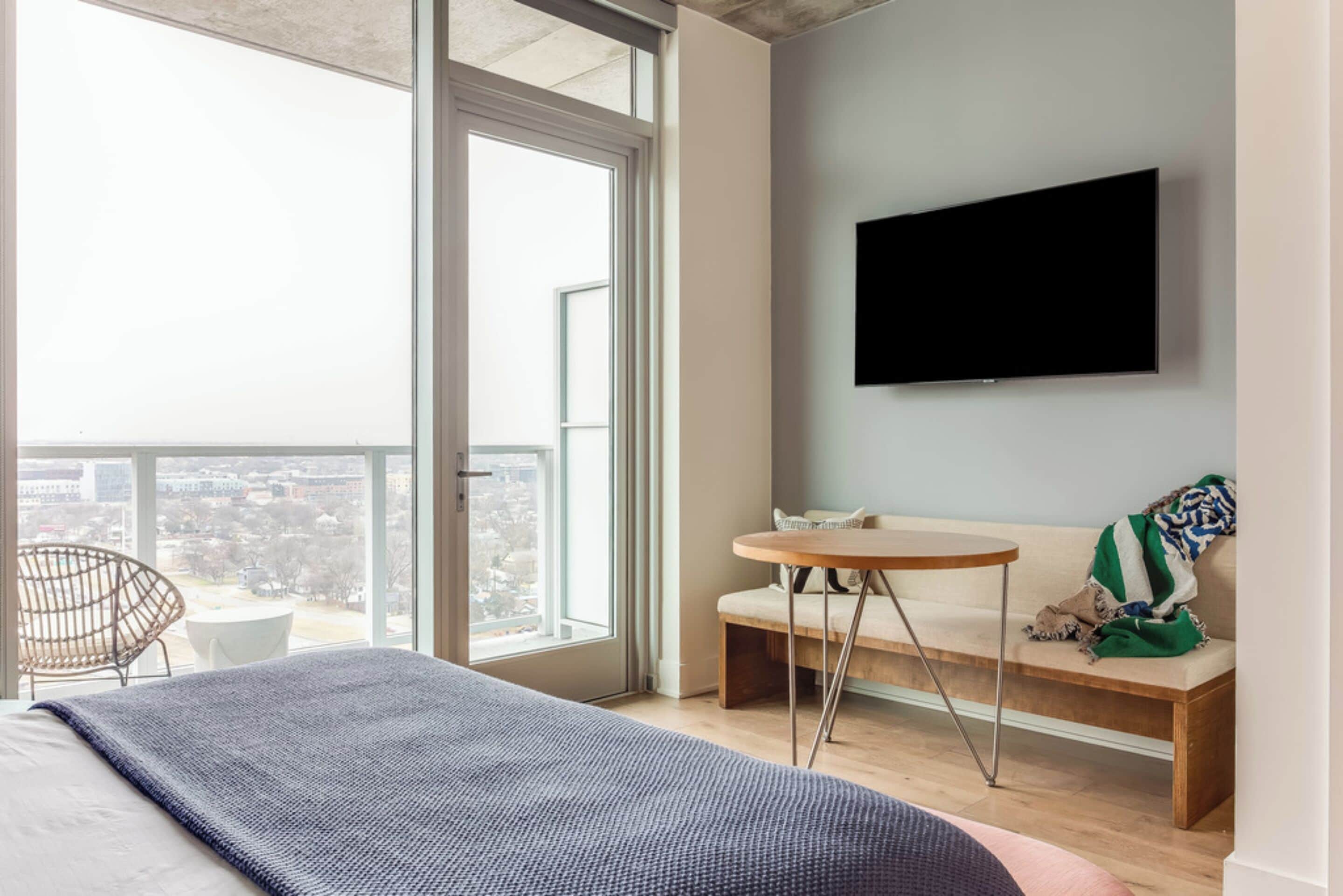 A modern bedroom with a large window, a bed, a round table, a sofa, and a TV mounted on the wall.