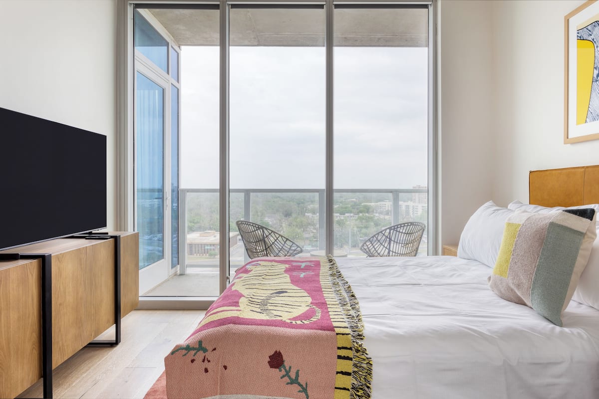 A modern bedroom with a large window, a bed with a colorful blanket, a TV, and a wooden desk.