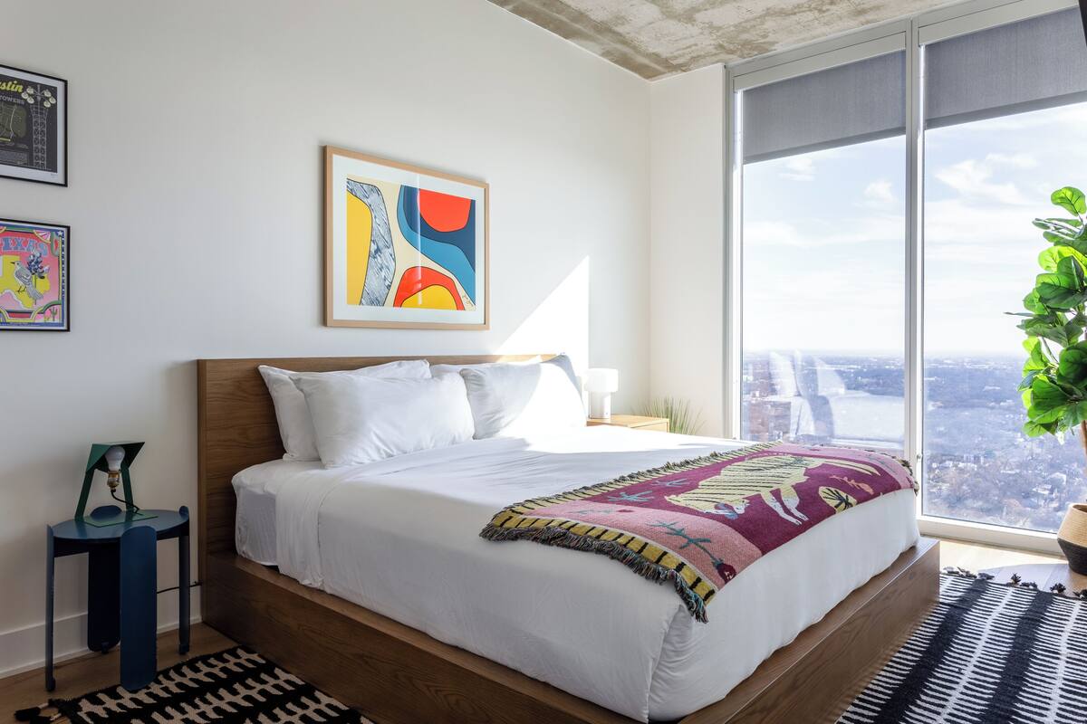 A modern bedroom with a large bed, a city view, and a framed abstract painting.