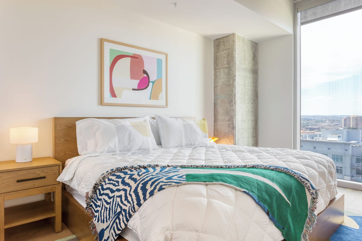 A hotel room with a large bed, a nightstand, a lamp, a colorful abstract painting, and a view of the city through a large window.