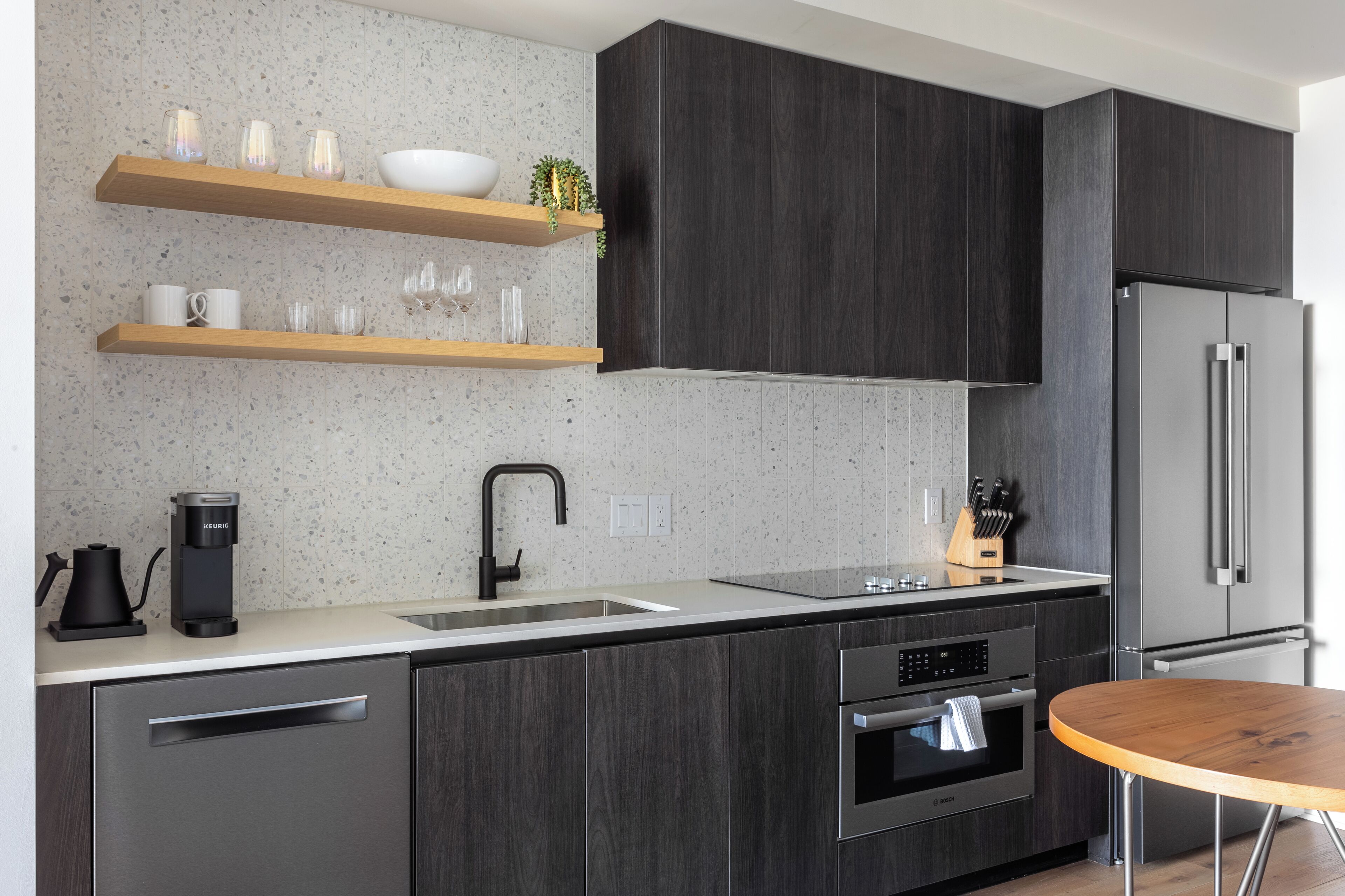 Une cuisine moderne avec des meubles de couleur bois foncé, un réfrigérateur en acier inoxydable, un four encastré et une table ronde en bois.