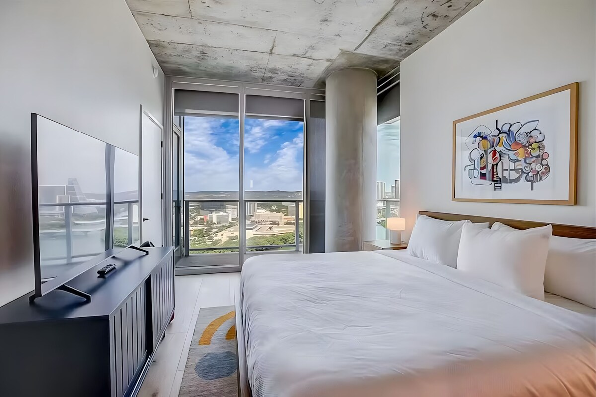 A modern hotel room with a large bed, a flat-screen TV, and a city view through large windows.