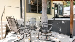 A covered outdoor seating area with metal chairs and a table, a tree, and a house in the background. - Magnolia Cottage | 3/2, Full Kitchen, Hot Tub, Grill, 3 Blocks to Main (Fredericksburg)