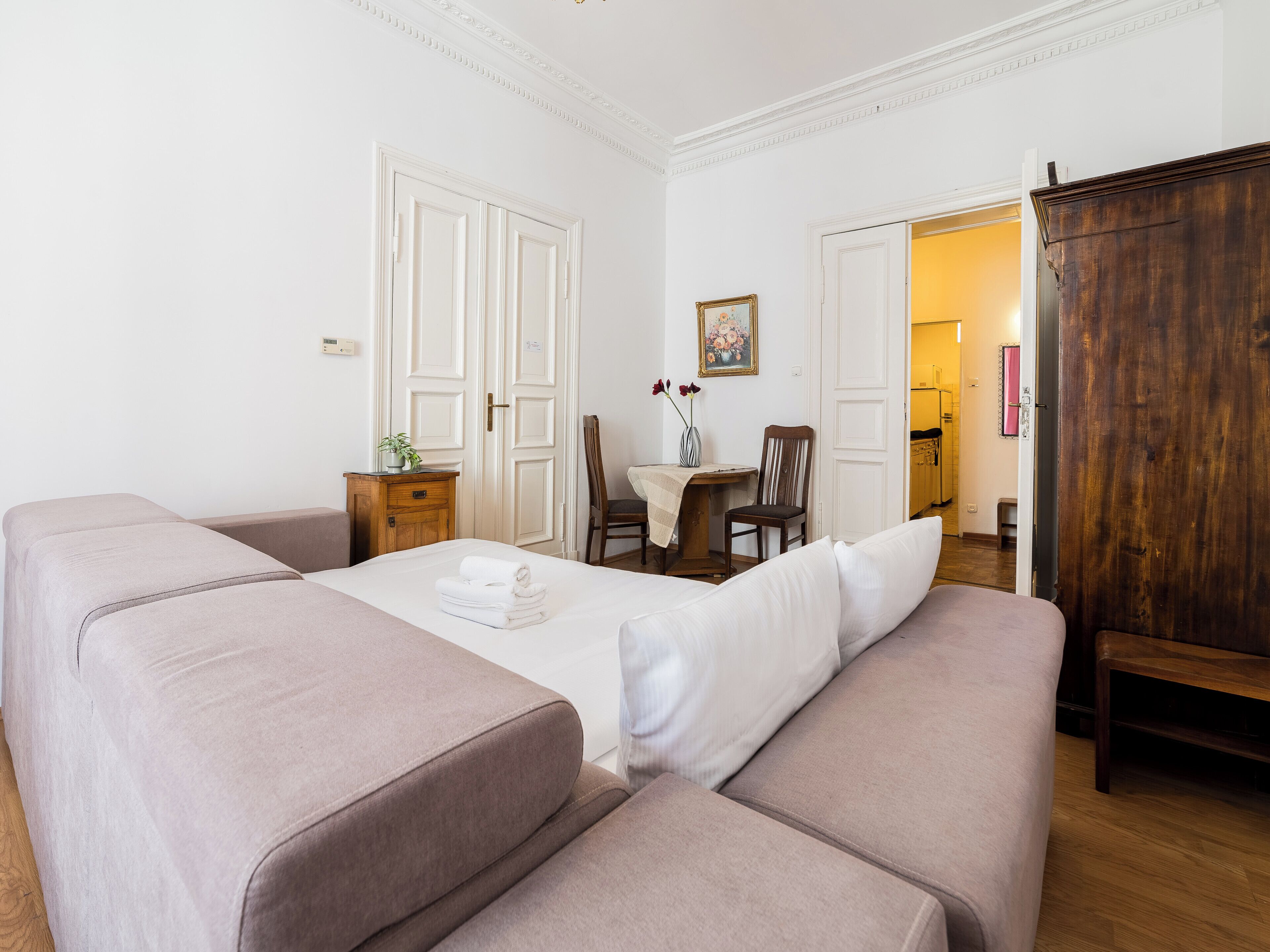 A room with a bed, a wooden wardrobe, a dining table with chairs, and a kitchen visible through an open door.