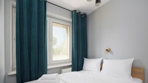 Una cama bien hecha con ropa blanca, una ventana con cortinas turquesa y una lámpara fijada a la pared.