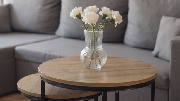 Una mesa redonda de madera con un jarrón de cristal transparente que contiene flores blancas, colocada sobre un sofá gris en una sala de estar.