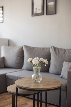 Een ronde houten tafel met een transparante glazen vaas met witte bloemen, geplaatst op een grijze bank in een woonkamer.