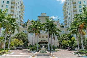 Um hotel moderno com uma fonte central, rodeado por palmeiras e várias varandas.