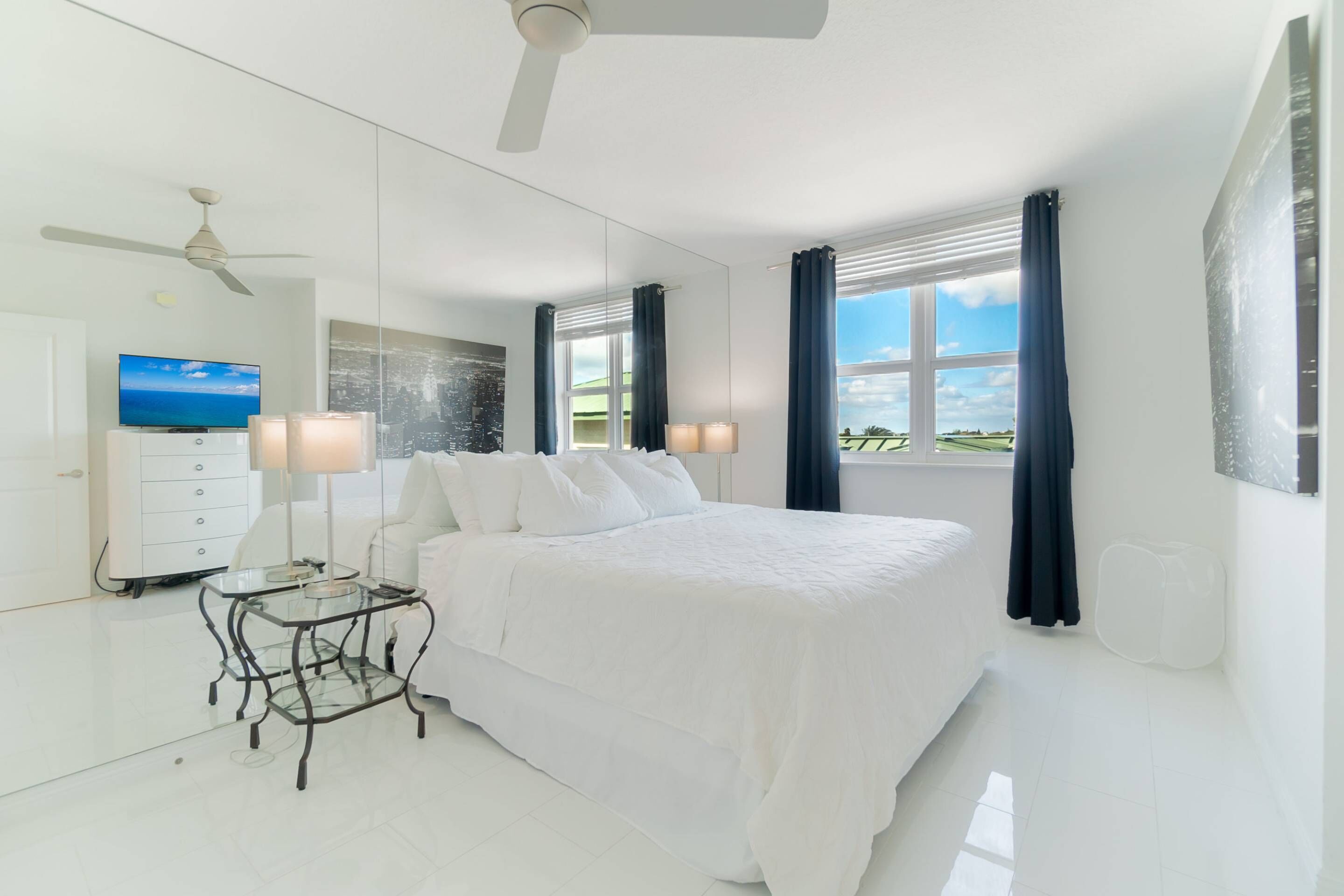 A modern bedroom with a large bed, a ceiling fan, a TV, and a view of the outdoors.
