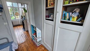Ein Zimmer mit Holzboden, einem Heizkörper, einem kleinen Tisch mit Lampe, einem Bücherregal mit Büchern und einem Stofftier, und einer Tür zu einem anderen Raum.