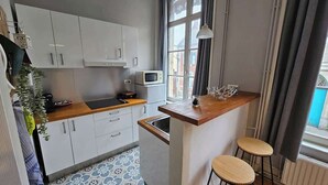 Eine kompakte Küche mit weißen Schränken, einer Mikrowelle, einem kleinen Kühlschrank, einer Holz Arbeitsplatte und einem Fenster mit Vorhängen.