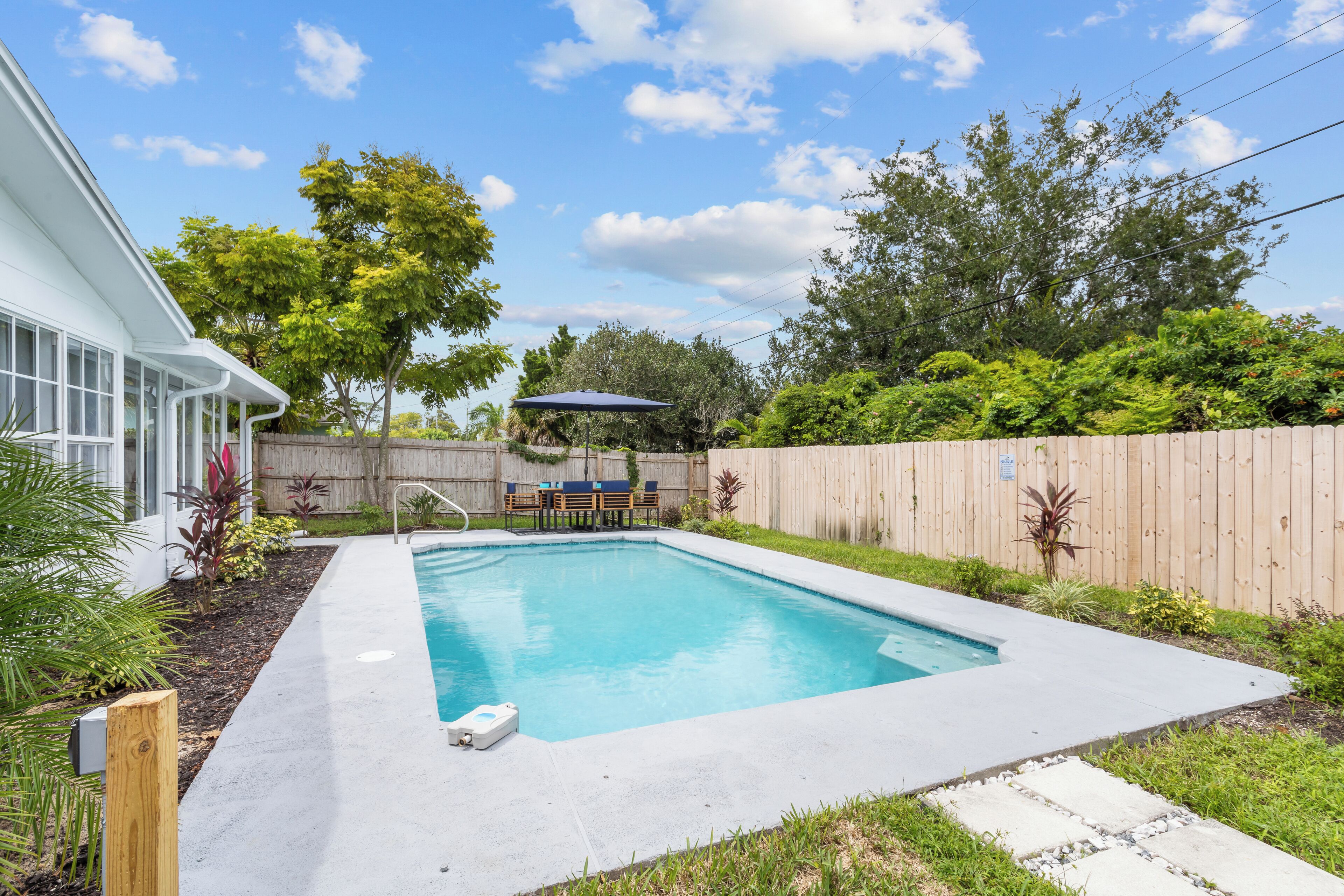A rectangular swimming pool with a wooden deck, surrounded by a wooden fence and lush greenery.