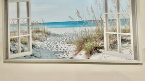 Une vue sur la plage à travers une fenêtre ouverte, avec un sentier de sable qui mène à l’eau.