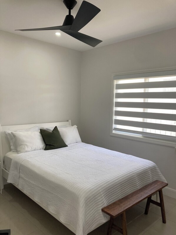 A bedroom with a bed, a bench, a ceiling fan, and a window with blinds. - Modern, Cozy, Airy 3-Bedroom Tropical Escape in Oceanpointe, Hanover, Jamaica (Lucea)