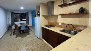 A compact kitchen with a gas stove, a sink, and wooden cabinets. There is a dining area with a table and chairs, and a refrigerator. - Casa Ik. A 5 min del aeropuerto, 8 min del Centro
 (Mérida)