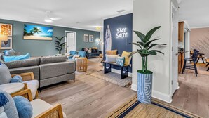 A modern living room with a grey sofa, a coffee table, and a TV displaying a beach scene. A wall with a 'Stay Salty' sign and a plant are also visible. - Near AMI-Heated Pool-Hot Tub-Game Room-Putting Green-Outdoor Games (Bradenton)
