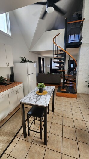 A modern kitchen with white cabinets, a marble countertop, and a dining area with a table and chairs. A spiral staircase leads to an upper level with a balcony. - Timeless Tremont (Cleveland)