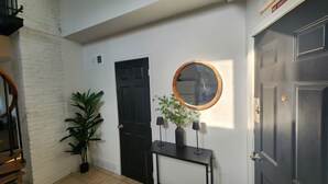 A hallway with a black door, a white wall, a wooden-framed mirror, a potted plant, and a small table with two lamps. - Timeless Tremont- Enjoy 20% off 5+ nights  (Cleveland)
