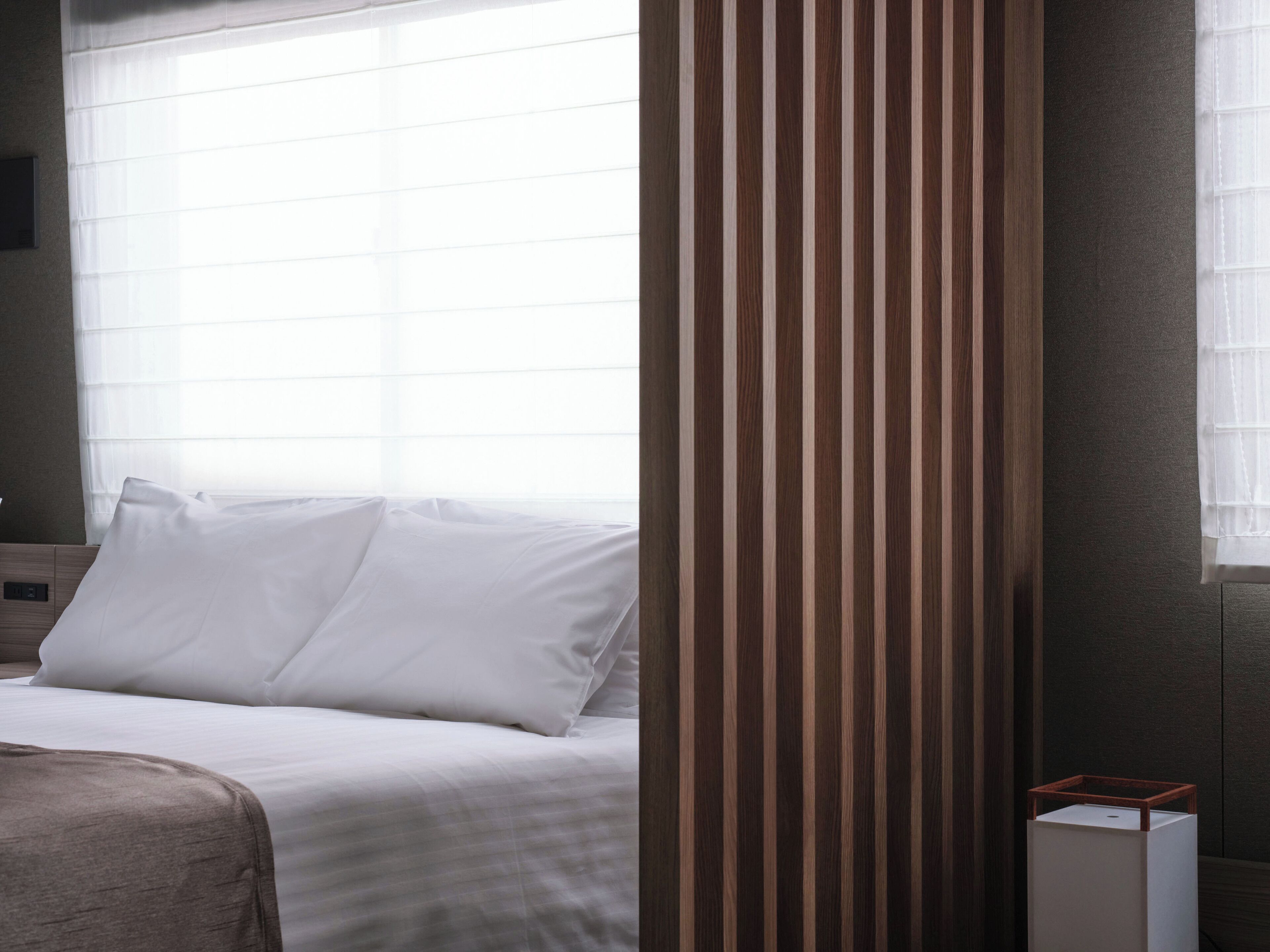 A hotel room with a bed, a window with blinds, a wooden panel, and a small white table with a wooden top.