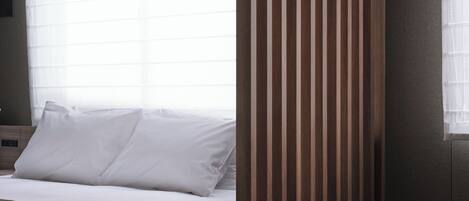 A hotel room with a bed, a window with blinds, a wooden panel, and a small white table with a wooden top.