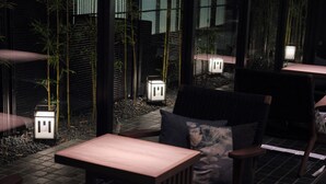 Un área de estar al aire libre tenuemente iluminada, con mesas y sillas, rodeada de plantas de bambú y un piso de baldosas.