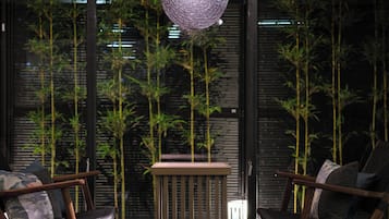 A modern interior with two wooden chairs, a small wooden table, and a hanging light fixture. Bamboo plants are visible through the window.