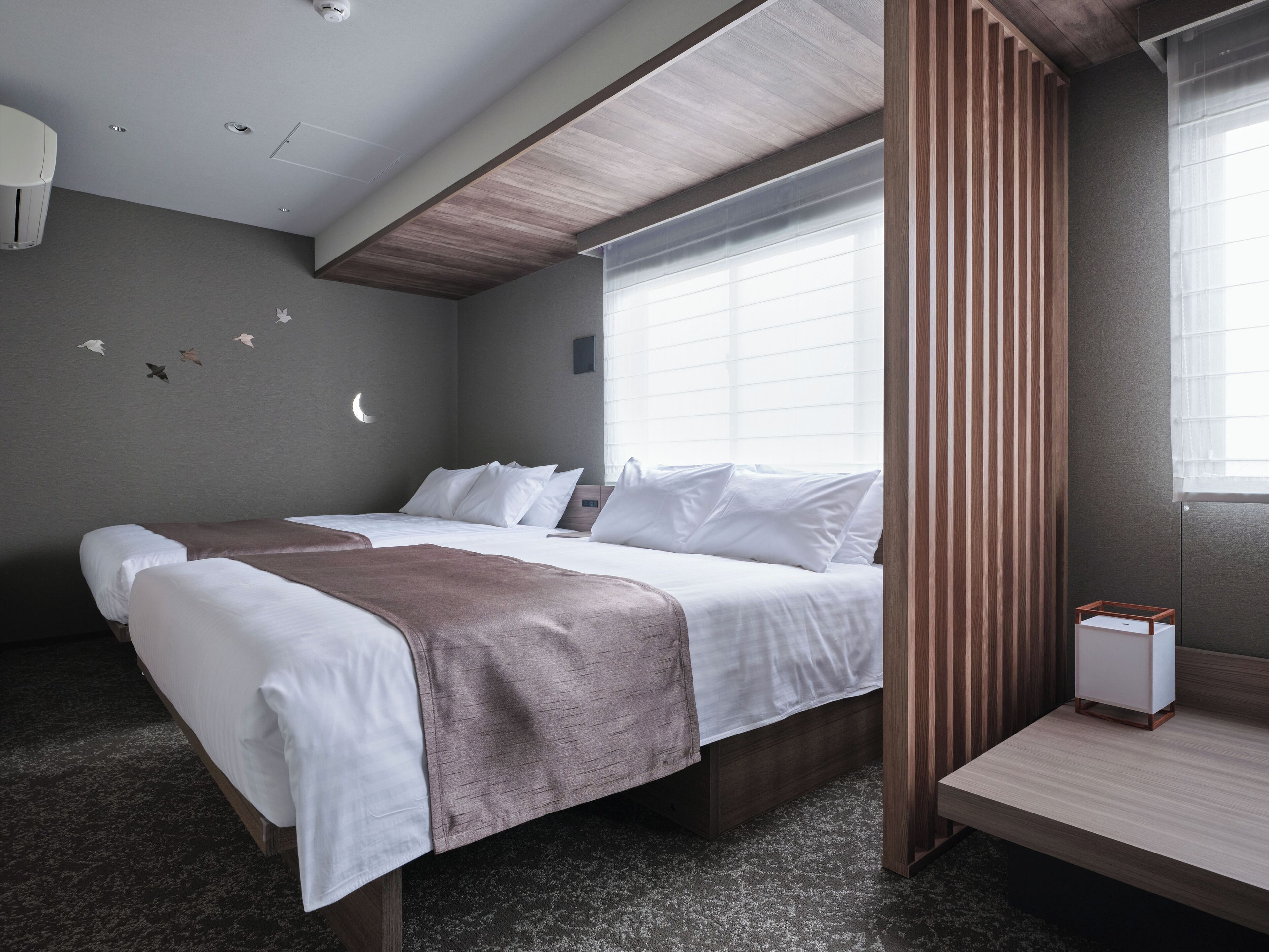 A hotel room with a bed, a bedside table, a window with blinds, a wall-mounted air purifier, and a ceiling light.