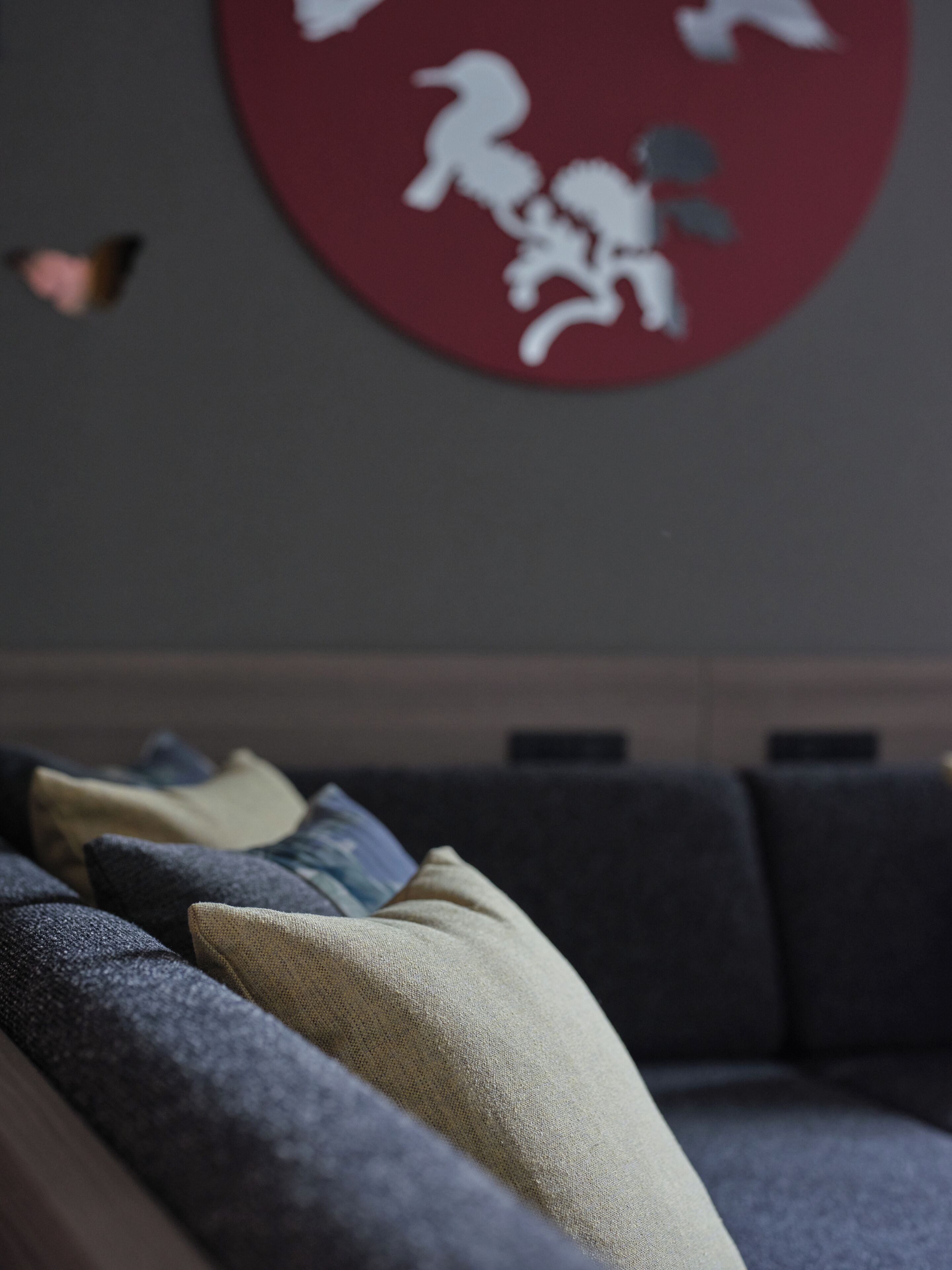 A living room with a dark grey sofa, beige and blue cushions, and a red circular wall decoration.