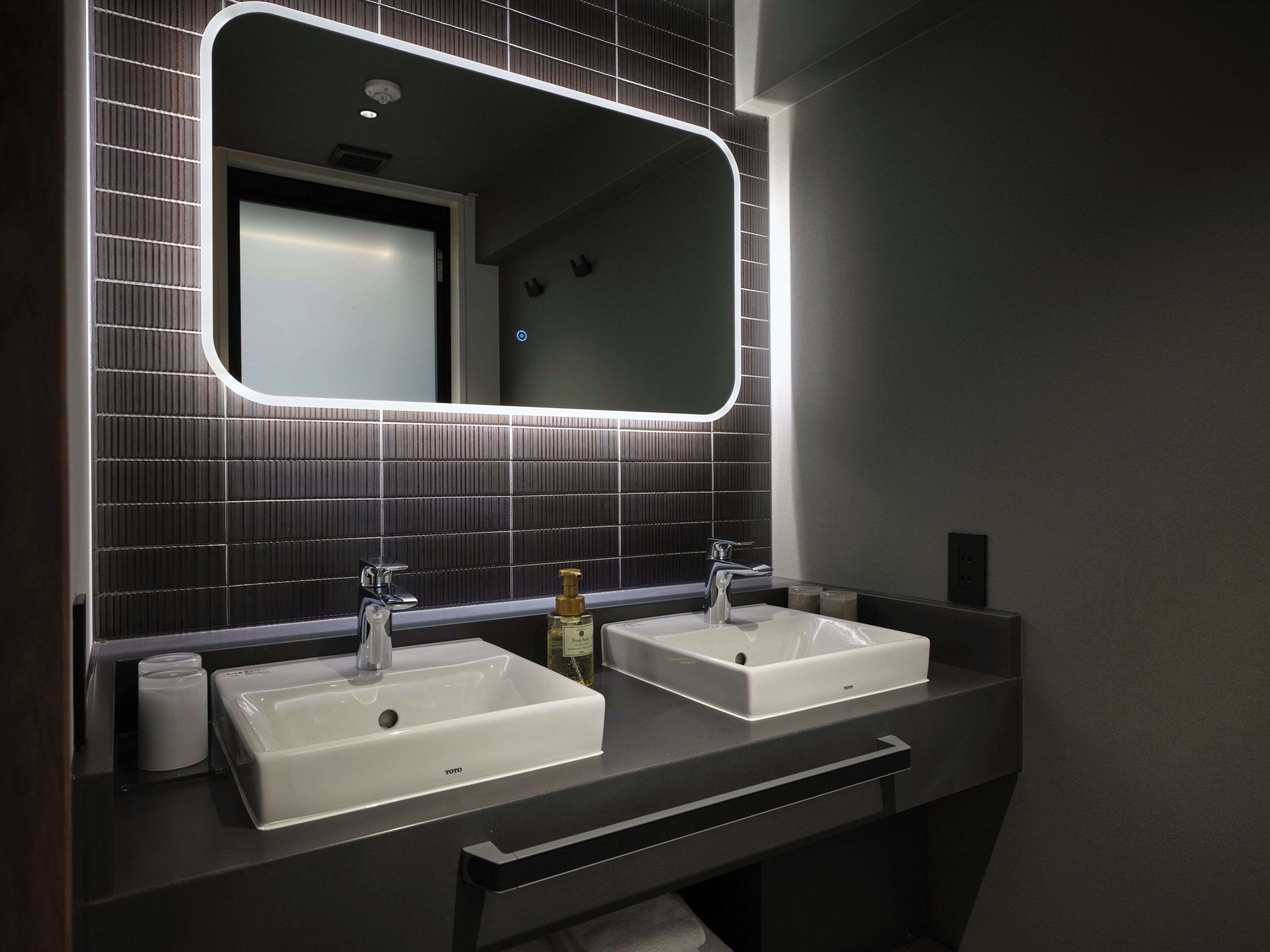 A modern bathroom with two white sinks, a large mirror with LED lighting, and a dark tiled backsplash.
