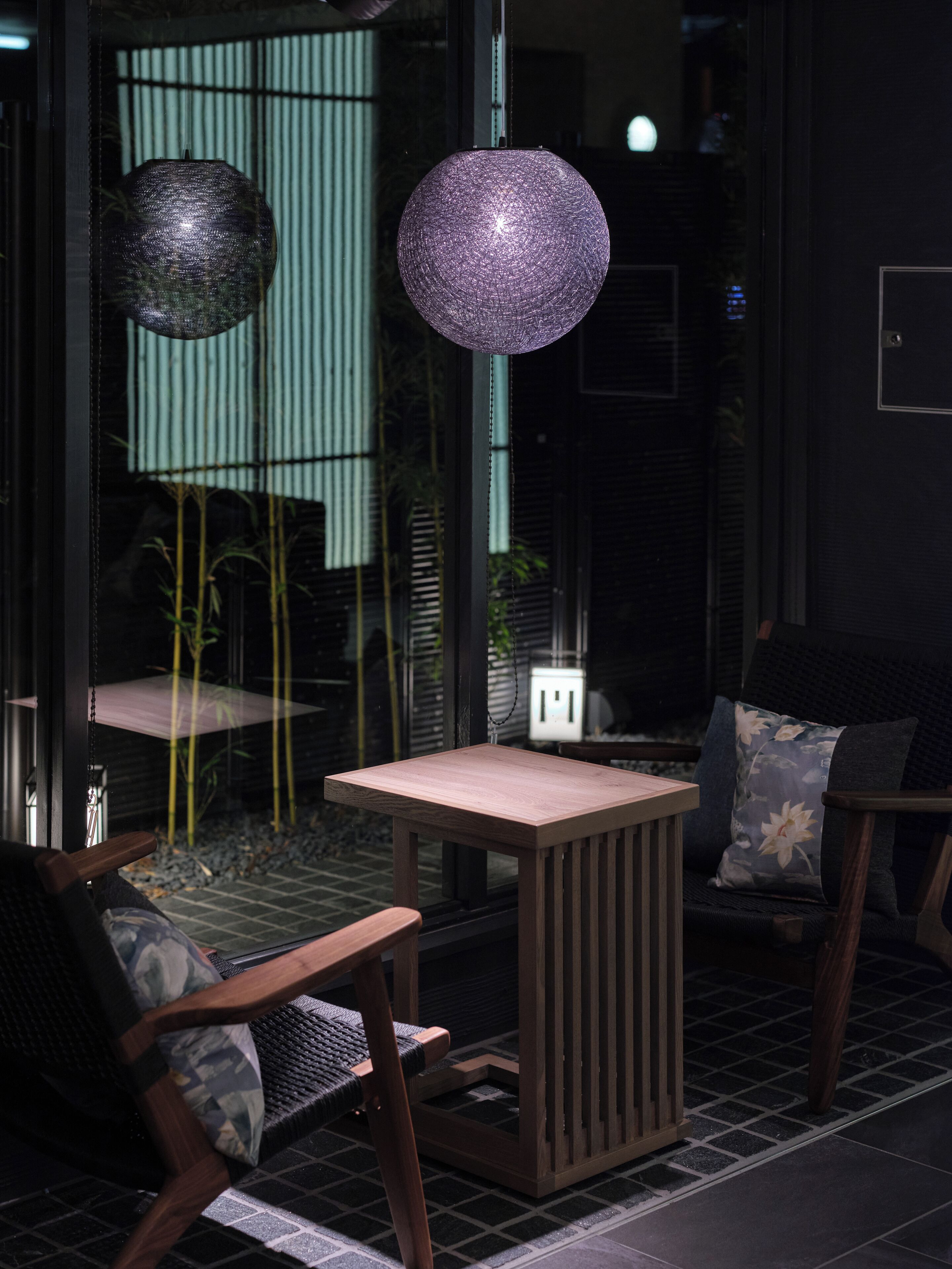 Un interior moderno con una mesa y sillas de madera, una lámpara colgante esférica y vistas a plantas de bambú a través de una puerta de cristal.
