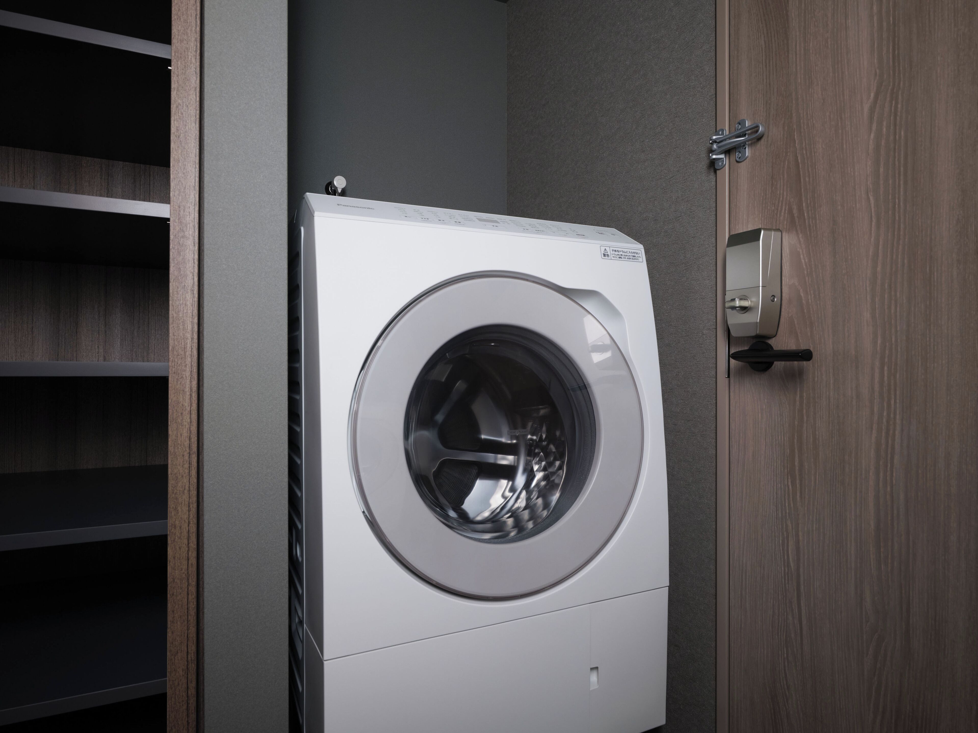 A front-loading washing machine in a laundry room with wooden shelves and a door.