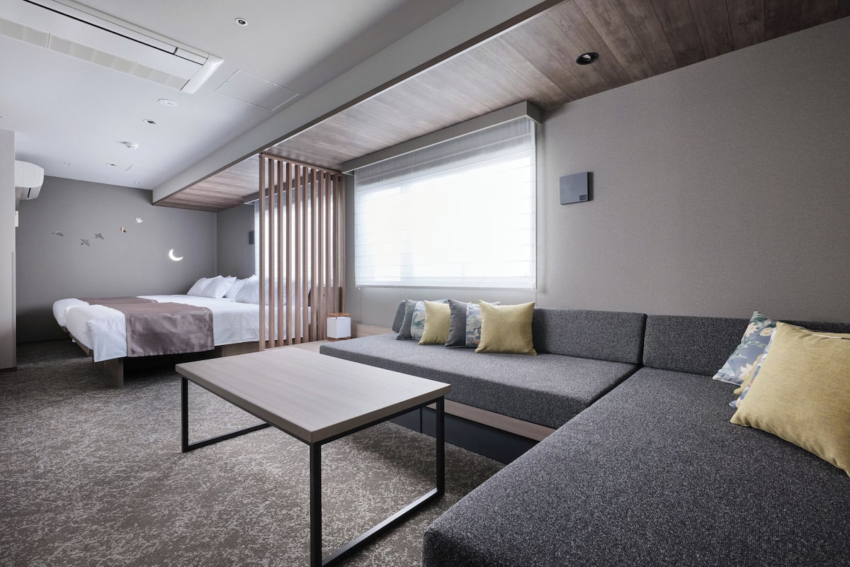 A modern hotel room with a bed, a sofa, a small table, and a window.