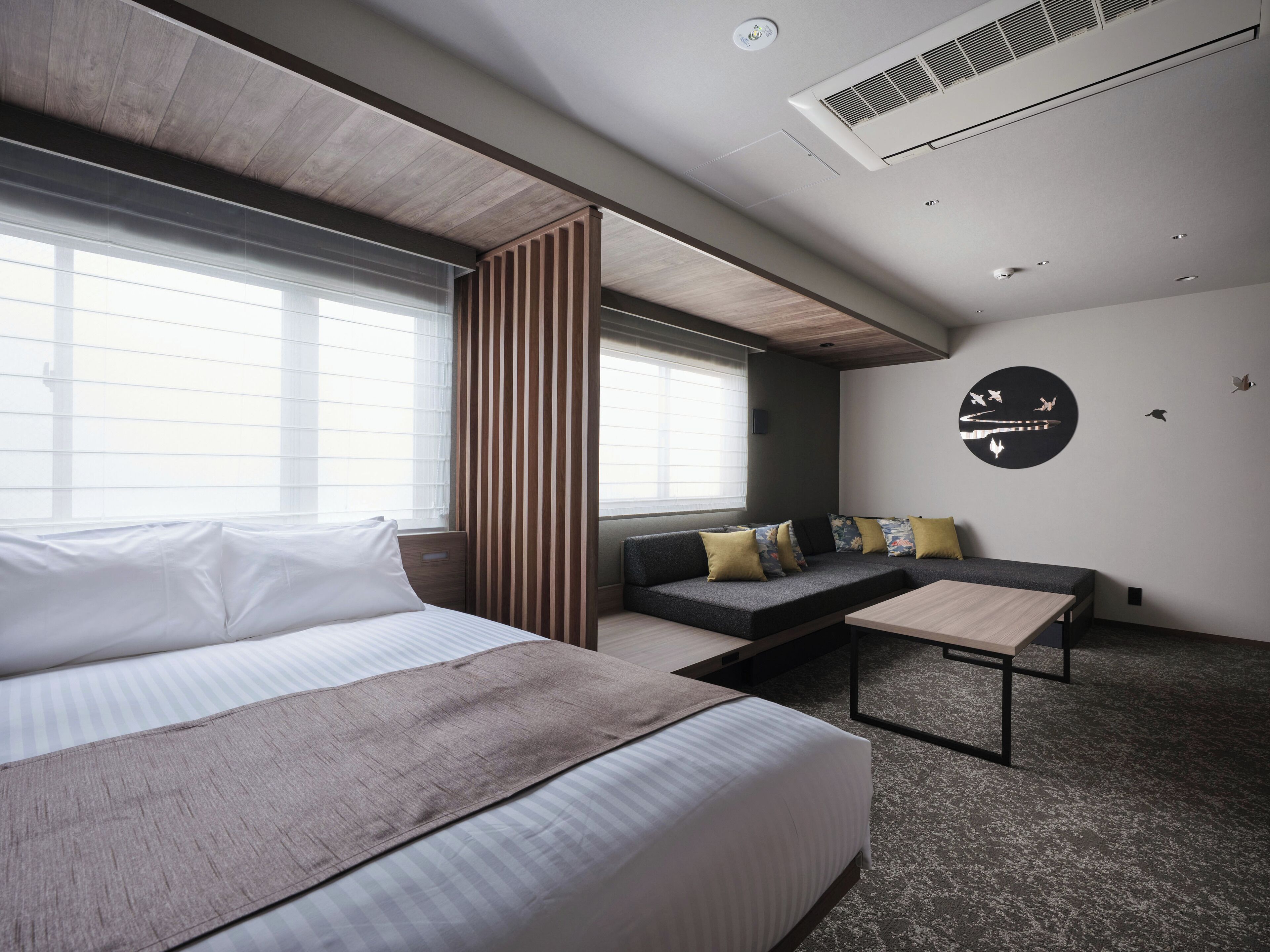A modern hotel room with a large bed, a sofa, a small table, and a wall-mounted air conditioner.