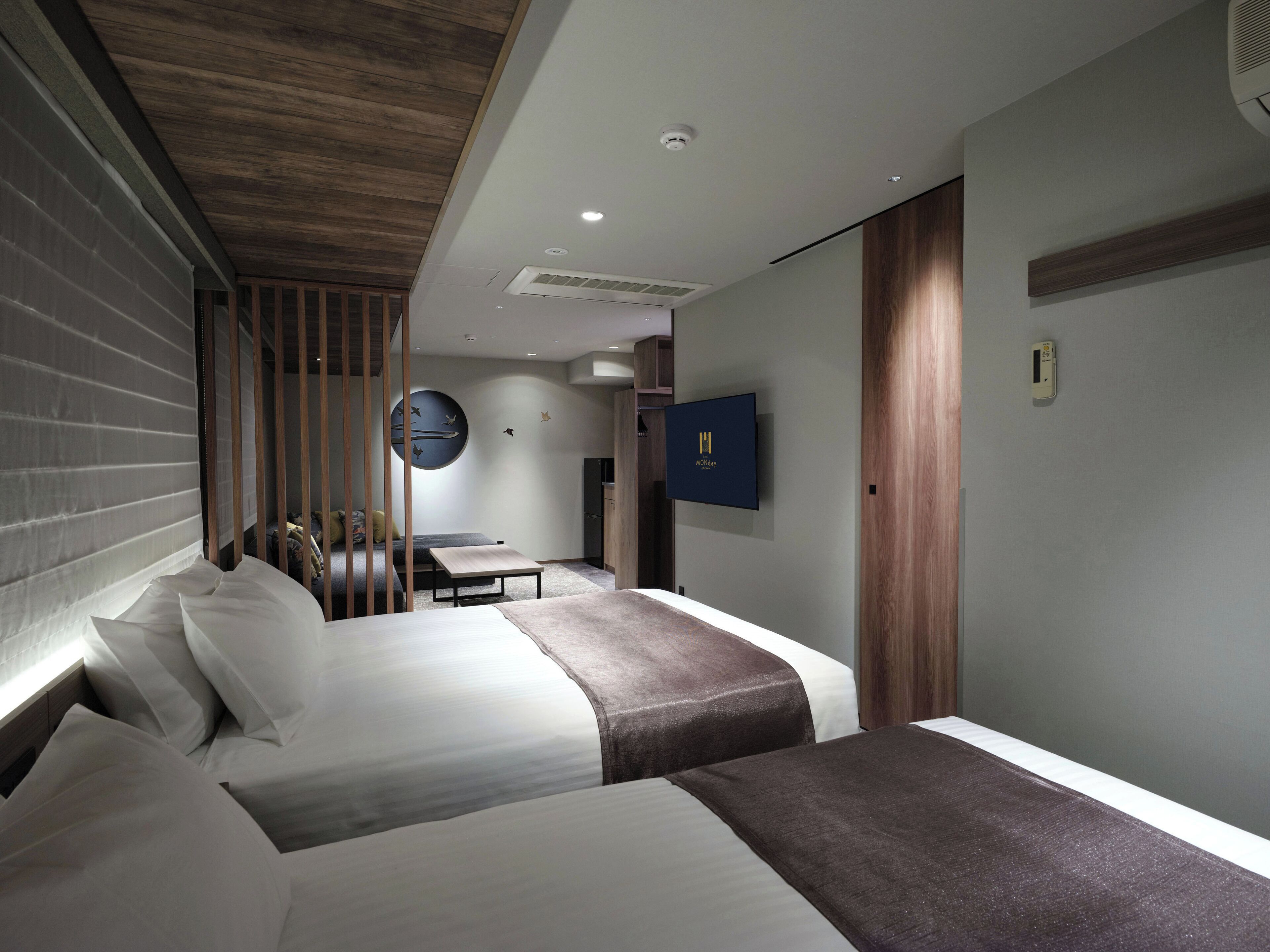 A hotel room with two beds, a desk, a TV, and a wall-mounted air conditioner.