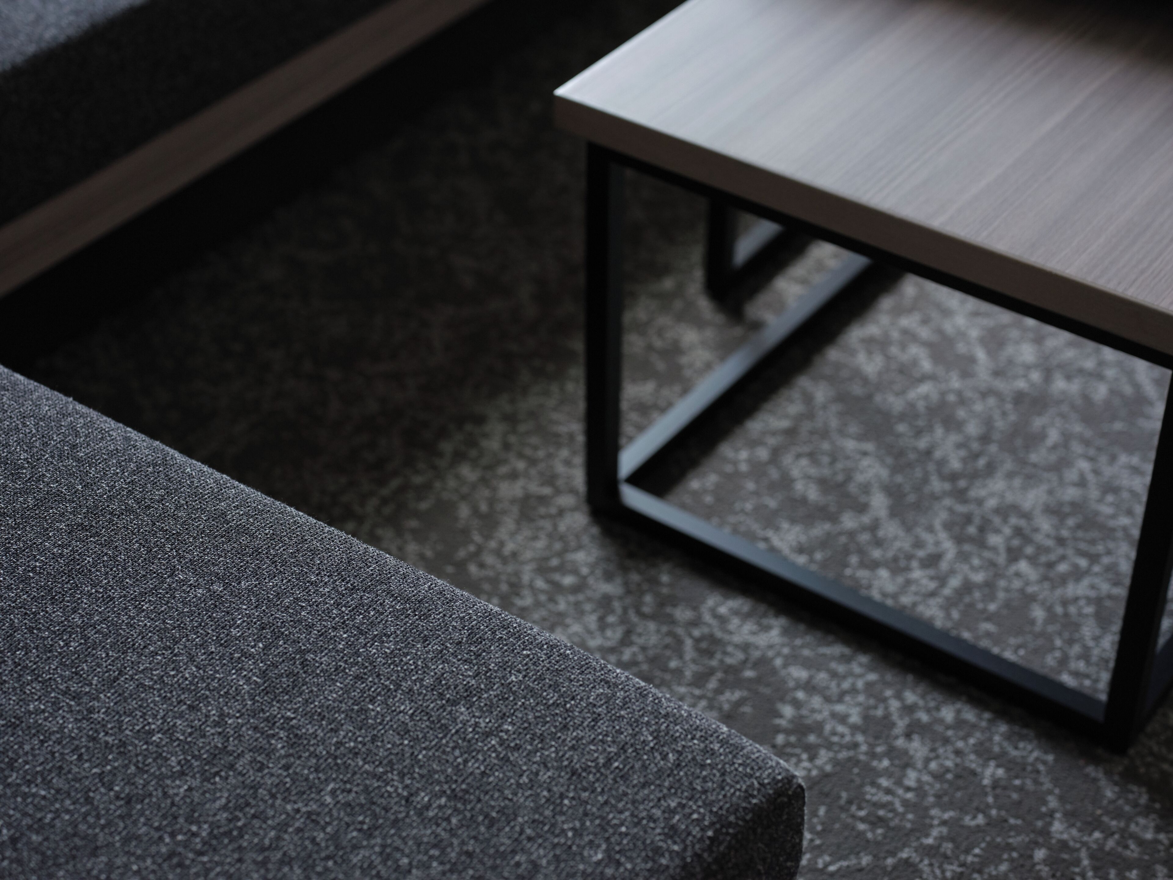 Habitació moderna amb un sofà gris fosc, una taula baixa de fusta amb estructura metàl·lica i una catifes gris amb relleu.