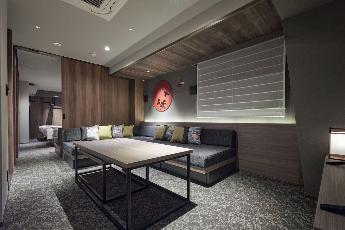 A modern living room with a sofa, a coffee table, and a dining table. There is a window with blinds, a wall-mounted air conditioner, and a ceiling light.