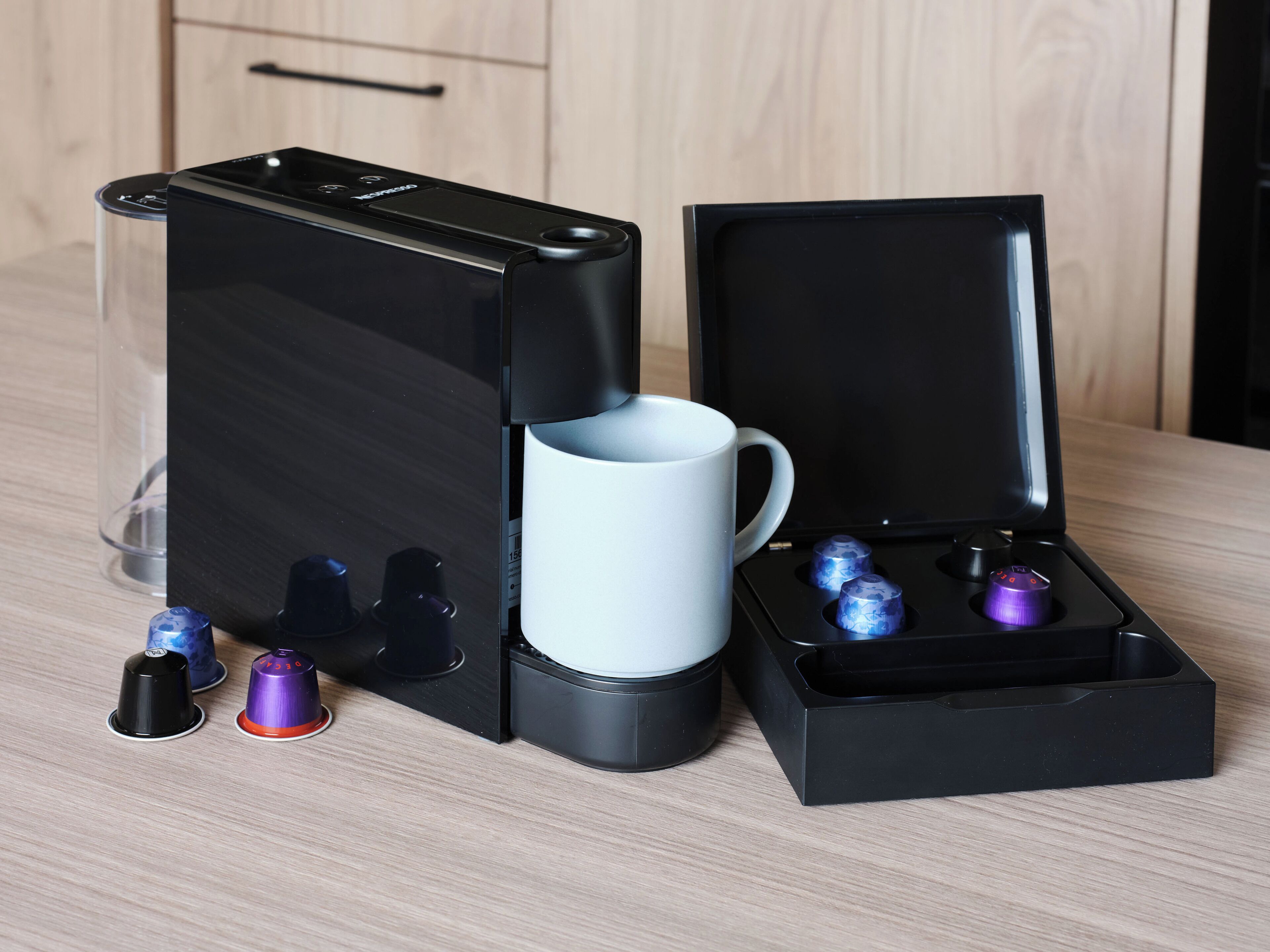 A coffee machine with a cup holder and a mug, and a box with two capsules.