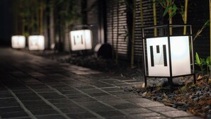 Un sendero bordeado por faroles iluminados y plantas de bambú, que conduce a un edificio con fachada de ladrillo.