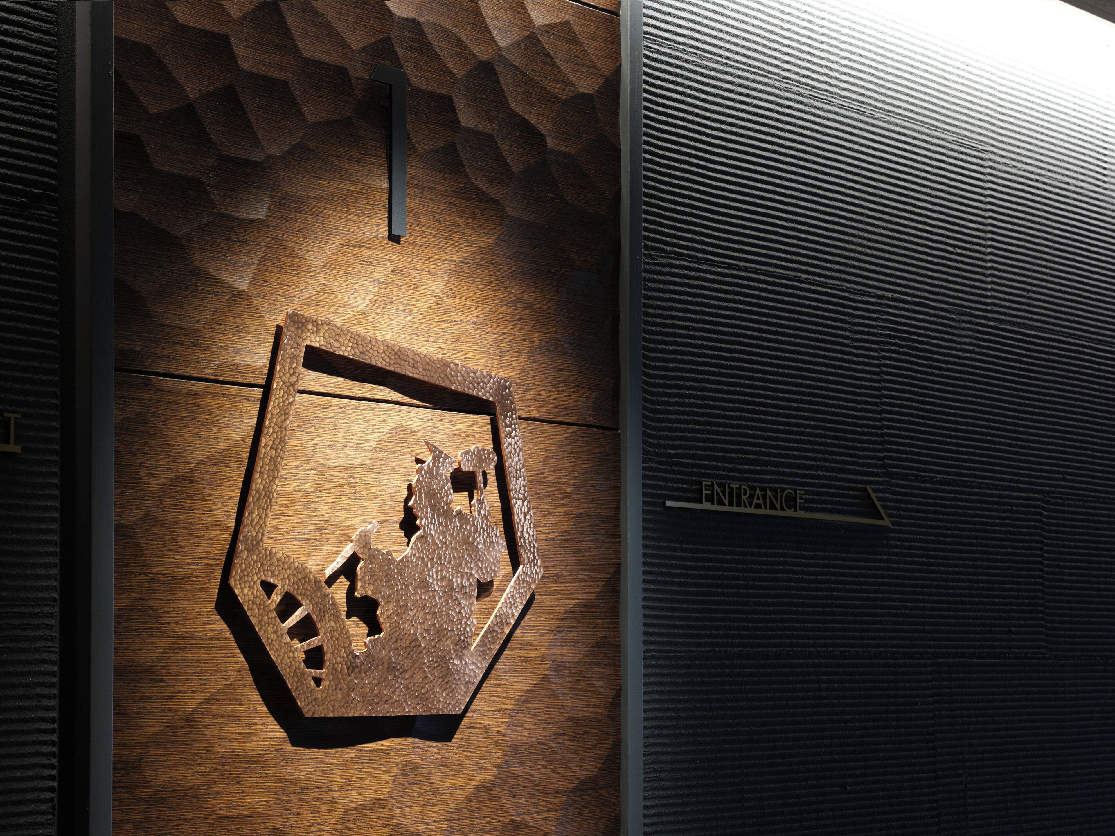 A hexagonal wooden sign with a textured surface and cut-out shapes, mounted on a wall with a ribbed panel to the right.