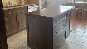 Una cocina con armarios de madera oscura, un lavamanos de acero inoxidable y una isla grande con cajones. Hay una ventana con cortinas y vistas al exterior.