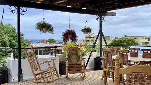 Una terraza de madera con mobiliario de mimbre, macetas colgantes y vistas al mar.