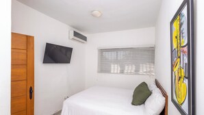 Un dormitorio con cama, puerta de madera, televisor montado en la pared, ventana con persianas y un cuadro enmarcado.