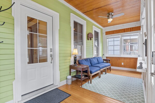 Fachada verde con techo de madera, puerta blanca, sofá azul y una pequeña mesa con lámpara.