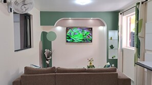 Een woonkamer met een bank, een aan de muur bevestigde televisie, een klein schap met decoratieve objecten en een raam met gordijnen.
