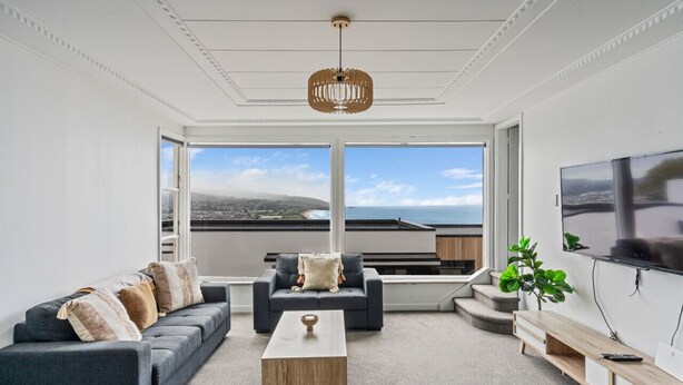A modern living room with a sofa, coffee table, and TV, large windows with a view of the city and ocean, and a ceiling light fixture. - St Clair Heights-Ocean views (Dunedin)