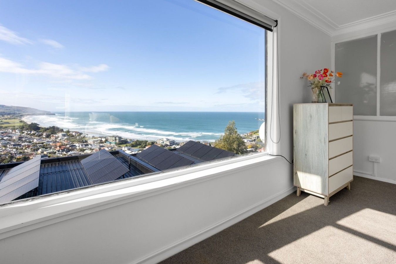A room with a large window overlooking a coastal town and ocean, a wooden dresser, and a vase with flowers.