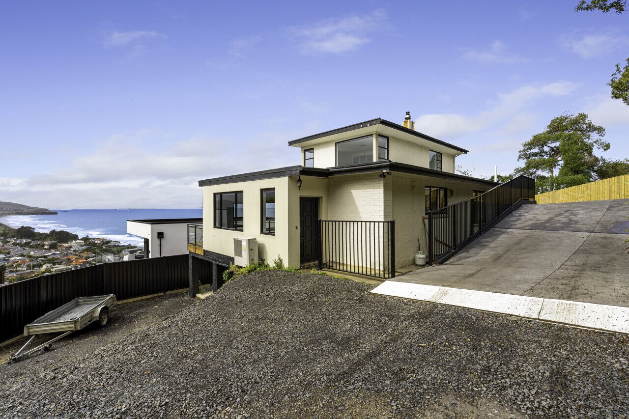 A modern two-story house with a view of the ocean, a gravel driveway, and a wooden fence.