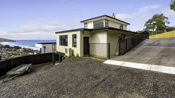 A modern two-story house with a view of the ocean, a gravel driveway, and a wooden fence. - St Clair Heights-Ocean views (Dunedin)