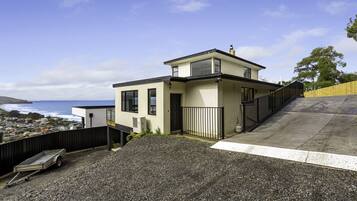 A modern two-story house with a view of the ocean, a gravel driveway, and a wooden fence.